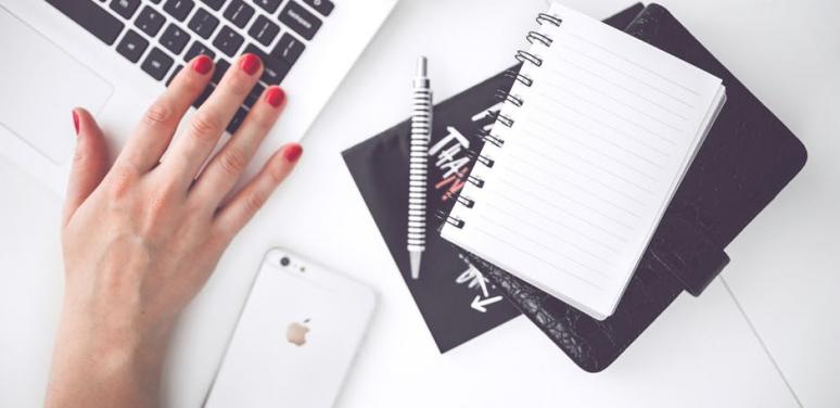 Image of a woman's hand on a laptop with a smart phone and a notebook and pencil next to it 